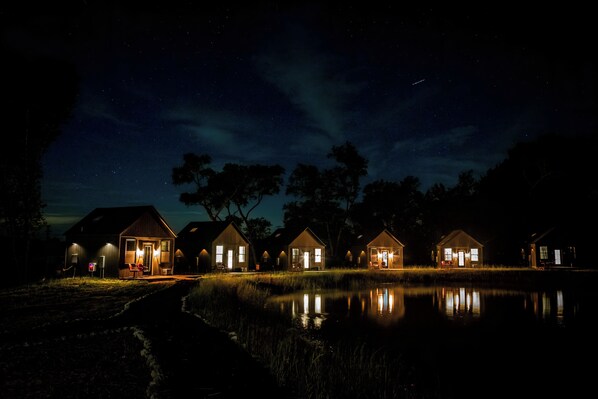 A row of cabins with illuminated windows and doors, situated by a body of water at night. - Limestone Fields (Jewett)