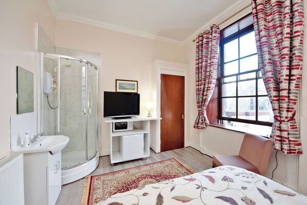 Habitación de hotel con ducha, televisor, mesita de noche, una silla y ventana con cortinas.