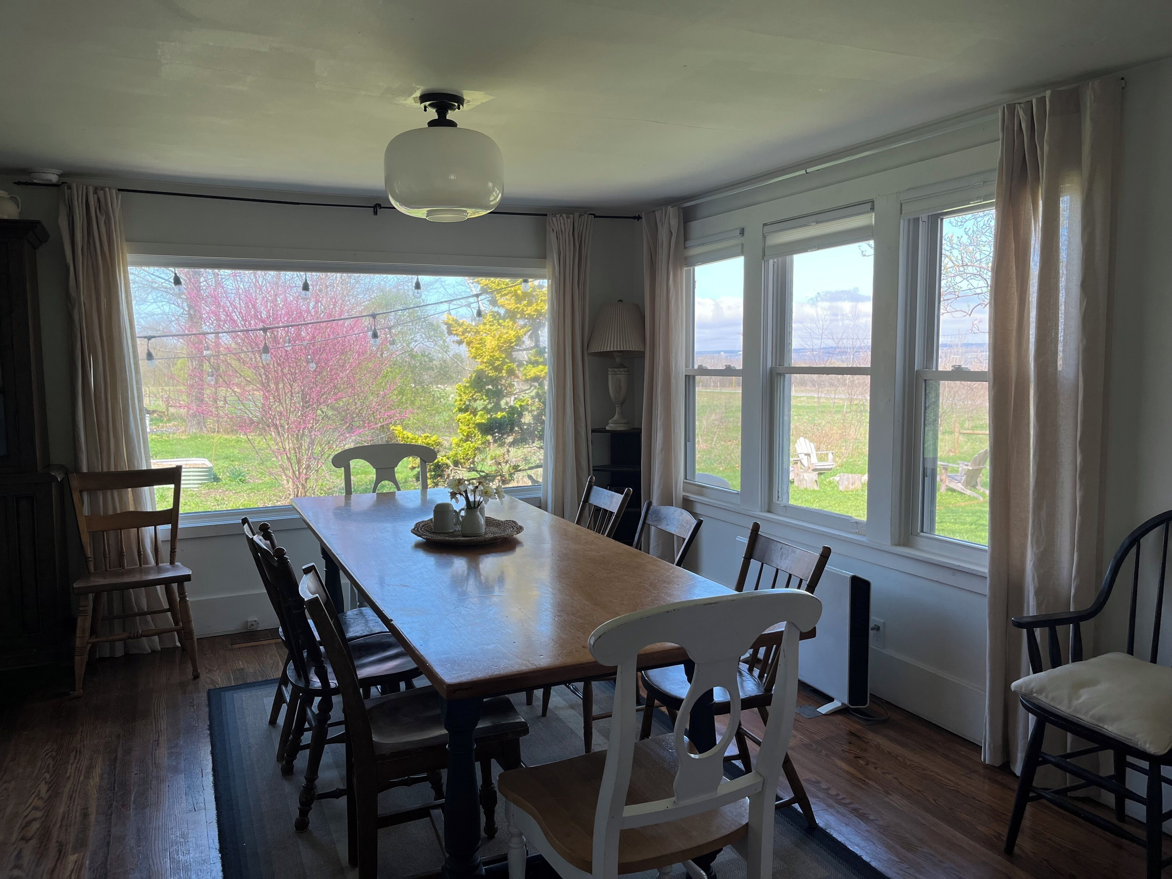 A dining room with a long wooden table, several chairs, a ceiling fan, and large windows with curtains.