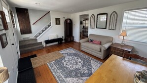 Una sala de estar con piso de madera, un sofá, una mesa de centro, una alfombra, un banco, una escalera y un comedor.