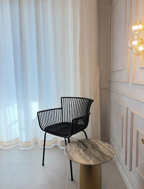 A chair with a unique design next to a round table in a room with a light-colored wall and a curtain.