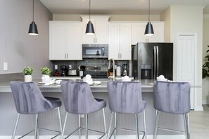 A modern kitchen with white cabinets, a stainless steel refrigerator, and four bar stools. - Magical 5BR Villa  Game Room & Pool  5mi to Disney (Kissimmee)