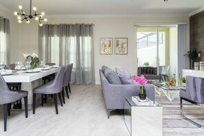 A modern living room with a grey sofa, a dining area with a white table, and a glass coffee table. - Magical 5BR Villa  Game Room & Pool  5mi to Disney (Kissimmee)