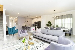 A modern living room with a grey sofa, blue armchair, and a coffee table. There is a dining area with a table and chairs in the background. - Magical 5BR Villa  Game Room & Pool  5mi to Disney (Kissimmee)