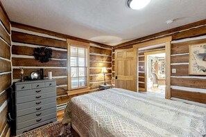 A log cabin-style bedroom with a bed, dresser, and a window with blinds. - Fisher Haul Historic Lodge | Hot Tub & Riverfront (South Fork)