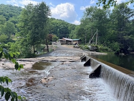A dam with a water spill over, surrounded by lush greenery and a building in the background.