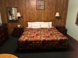 Quarto de hotel com parede de painéis de madeira, cama com colcha estampada, duas luminárias de cabeceira, mesa de cabeceira com telefone e pequena mesa com abajur.