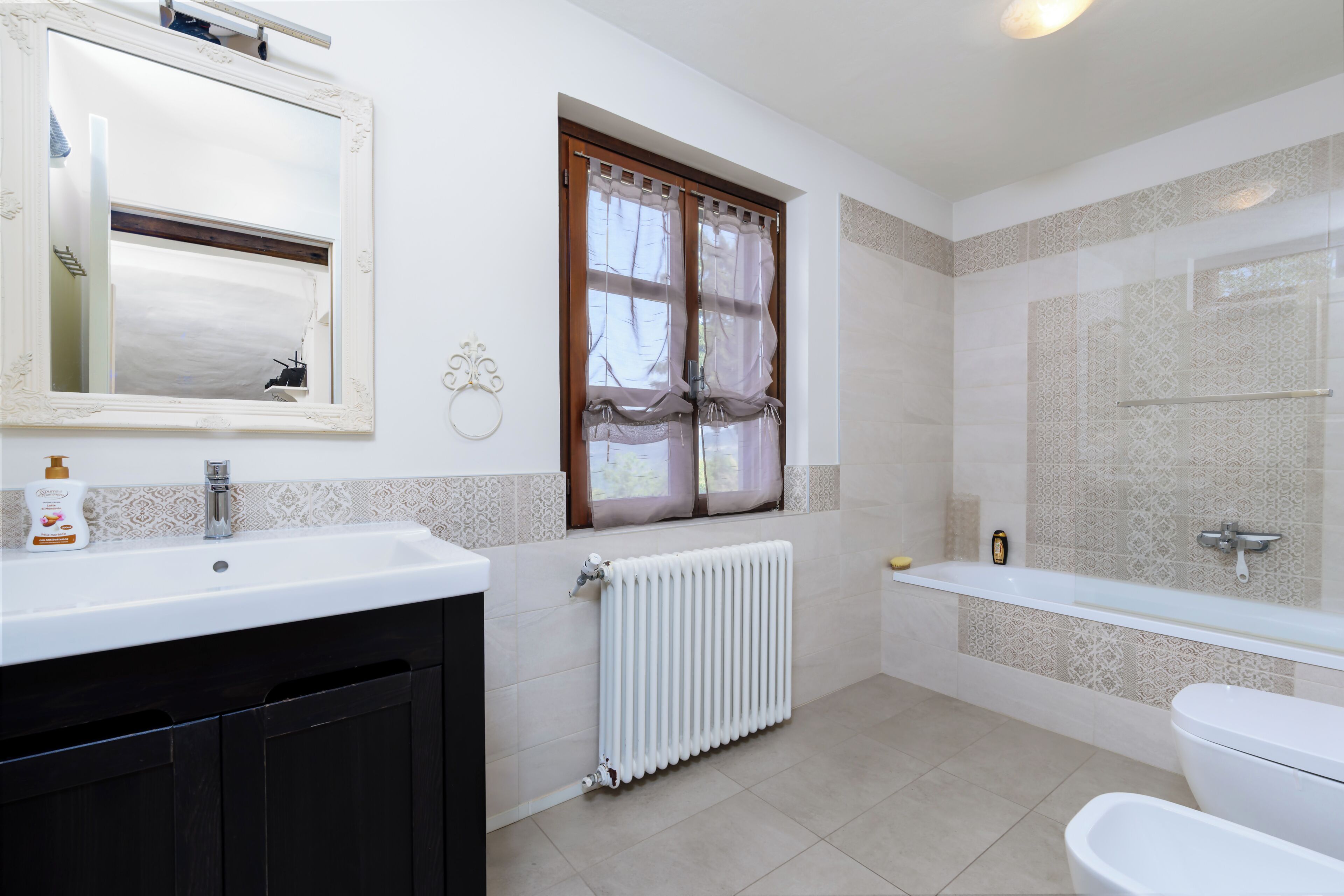 Une salle de bain avec un lavabo blanc, des toilettes, une baignoire, une fenêtre avec des rideaux et un radiateur.