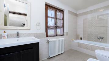 Une salle de bain avec un lavabo blanc, des toilettes, une baignoire, une fenêtre avec des rideaux et un radiateur.
