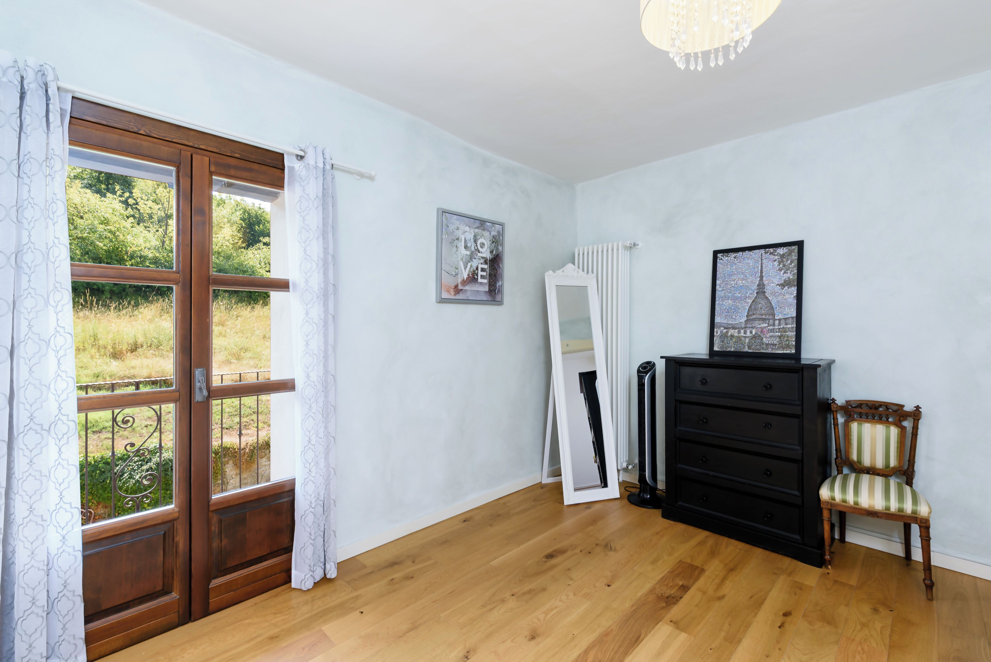 Ein Zimmer mit Holzboden, einem schwarzen Kommoden, einem Stuhl, einem Heizkörper und einem Fenster mit Vorhängen.