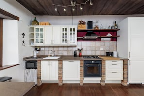 Una cocina con armarios blancos, una pared de acento de ladrillo, una estufa a gas y un estante rojo con utensilios de cocina.