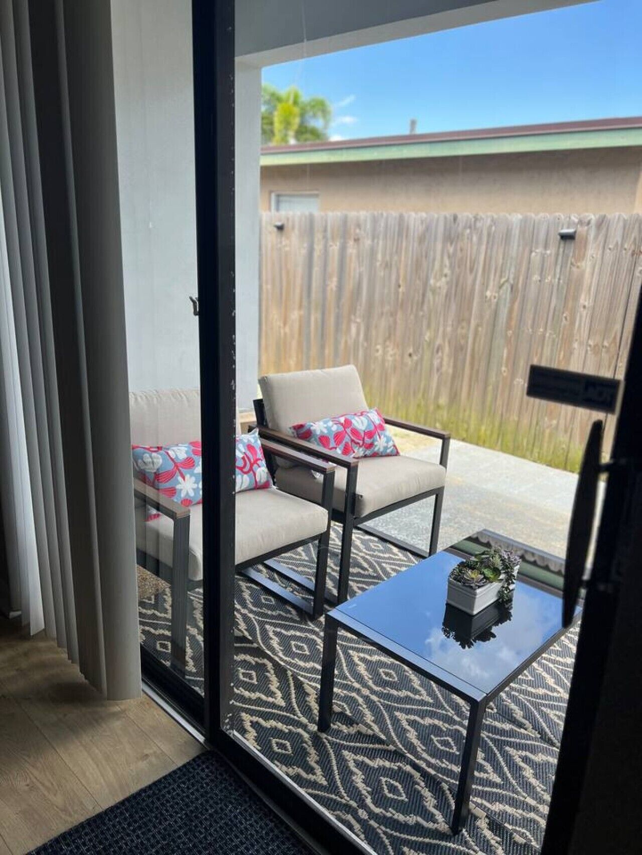 Una habitación con puerta corredera de vidrio que da a un patio al aire libre con cerramiento de madera, una silla acolchada, una mesita con una planta y un tapete estampado.