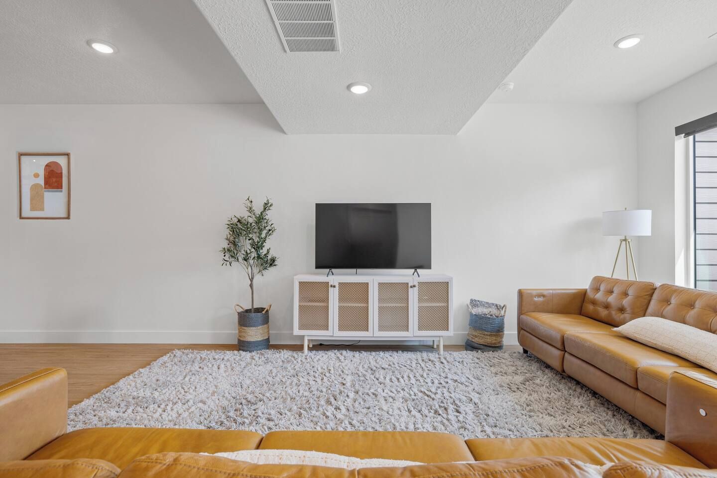 A modern living room with a flat-screen TV, a white cabinet, a brown leather sofa, and a large white rug.