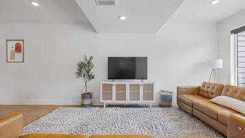 A modern living room with a flat-screen TV, a white cabinet, a brown leather sofa, and a large white rug.