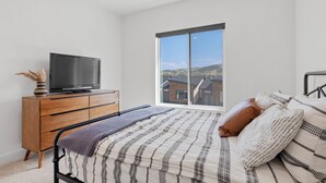 A bedroom with a bed, a dresser, a television, and a large window with a view of buildings. - Mountain Escape - Minutes to Snowbasin & Pineview - Sleeps 8 (Mountain Green)