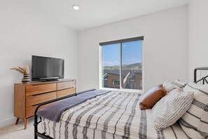 Ein Schlafzimmer mit einem Bett, einem Schminktisch, einem Fernseher und einem großen Fenster mit Blick auf Gebäude.