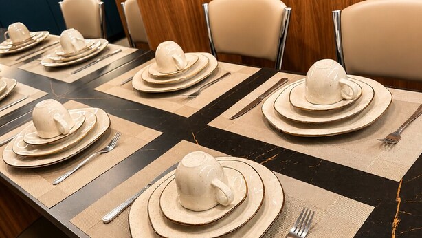 A dining table set with plates, bowls, and cutlery, with a wooden panel and chairs in the background. - Apartment with furnished balcony | Gym | Swimming pool | Complete linens (Goiânia)