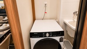 A compact laundry room with a front-loading washing machine, a small trash bin, and a sink. - Apartment with furnished balcony | Gym | Swimming pool | Complete linens (Goiânia)