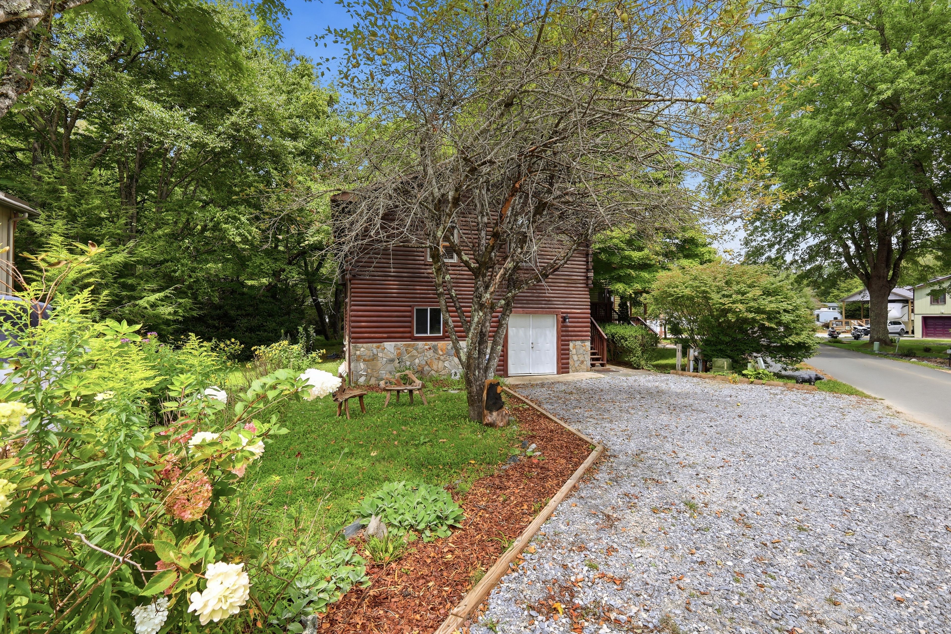 A log cabin with a stone foundation, surrounded by a gravel driveway and a garden with various plants and flowers.
