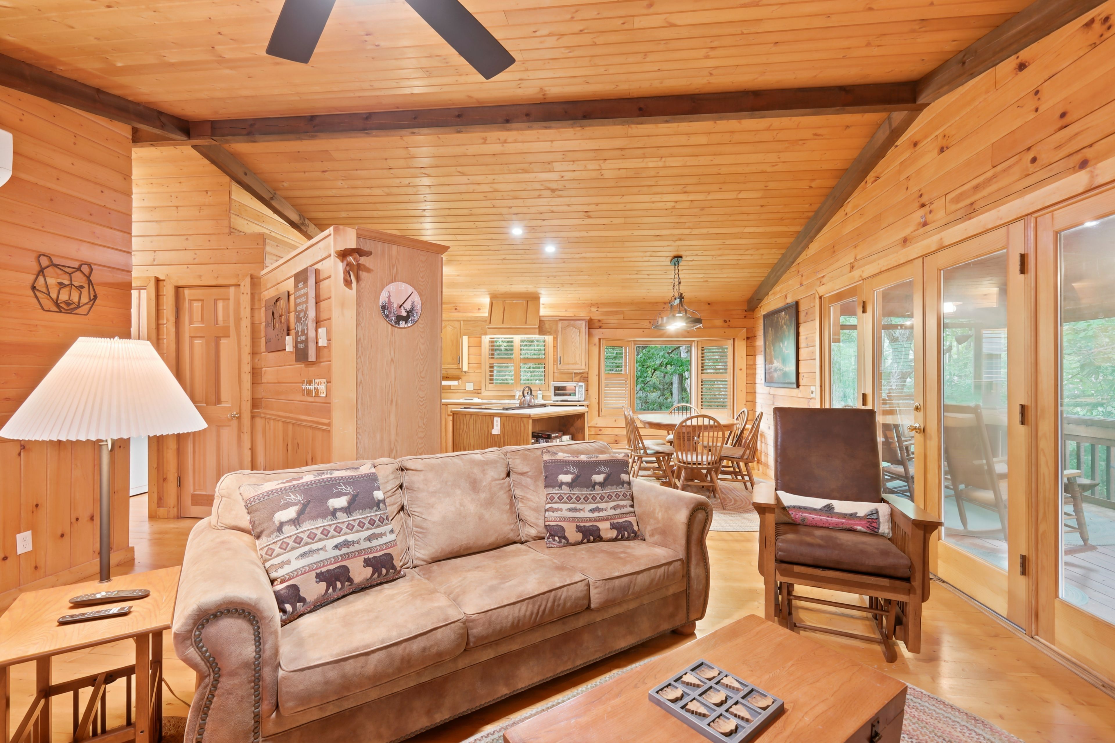 A cozy cabin interior with a wooden ceiling, a sofa with patterned cushions, a dining table with chairs, and a kitchen area.