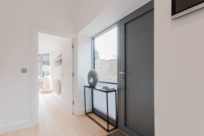 Een moderne hal met een glazen deur die uitkomt op een buitenruimte, een klein tafeltje met een kussen en een ingelijst beeld aan de muur.