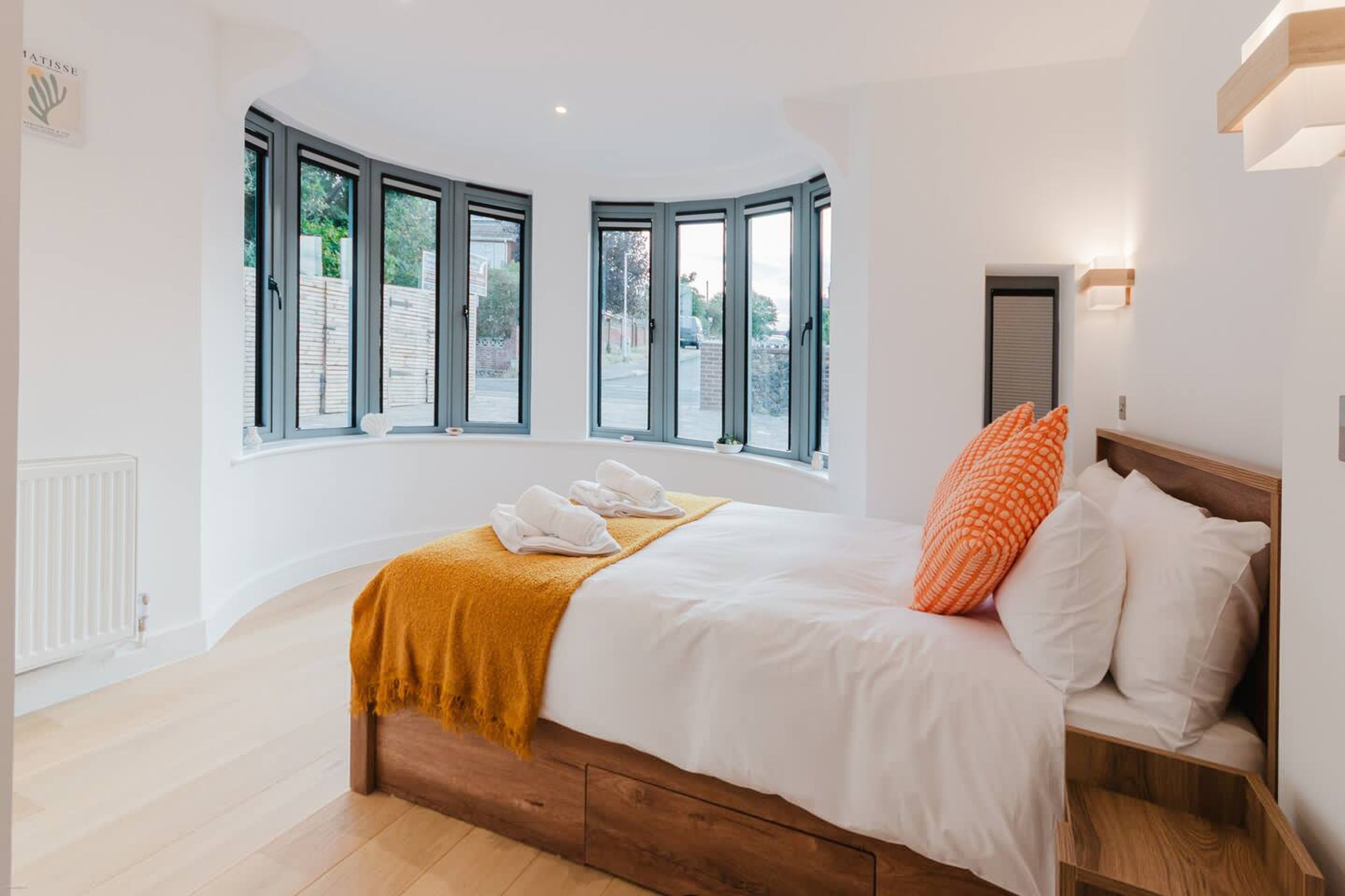 A bedroom with a large bed, a wooden headboard, a yellow throw blanket, and a view of the outside through large windows.