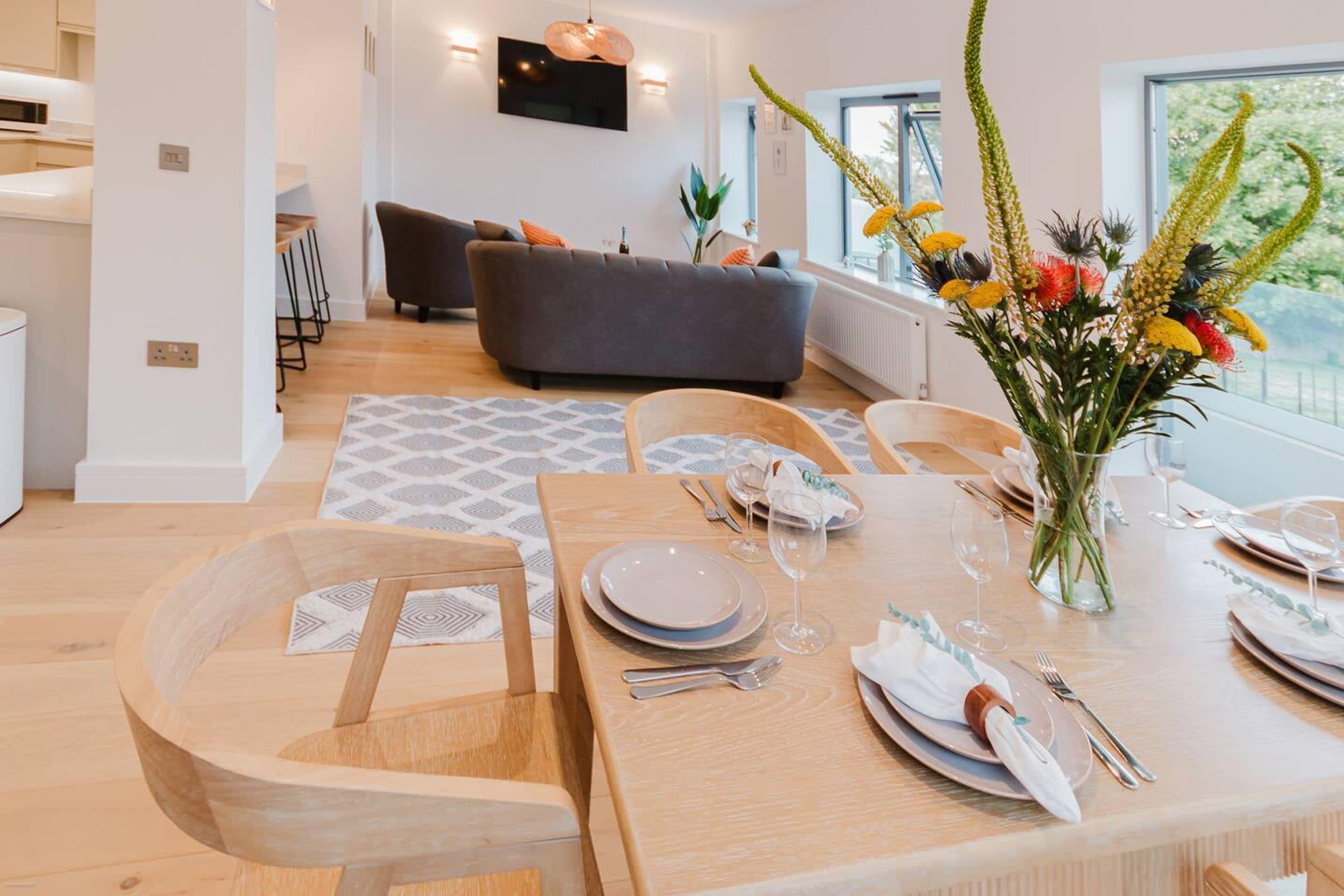 Een eetgedeelte met een houten tafel voor vier personen, een grijze bank op de achtergrond en een groot raam met uitzicht.