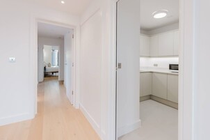 Un pasillo que conduce a un dormitorio con cama y ventana, una cocina con armarios blancos y microondas, y una puerta con pomo.
