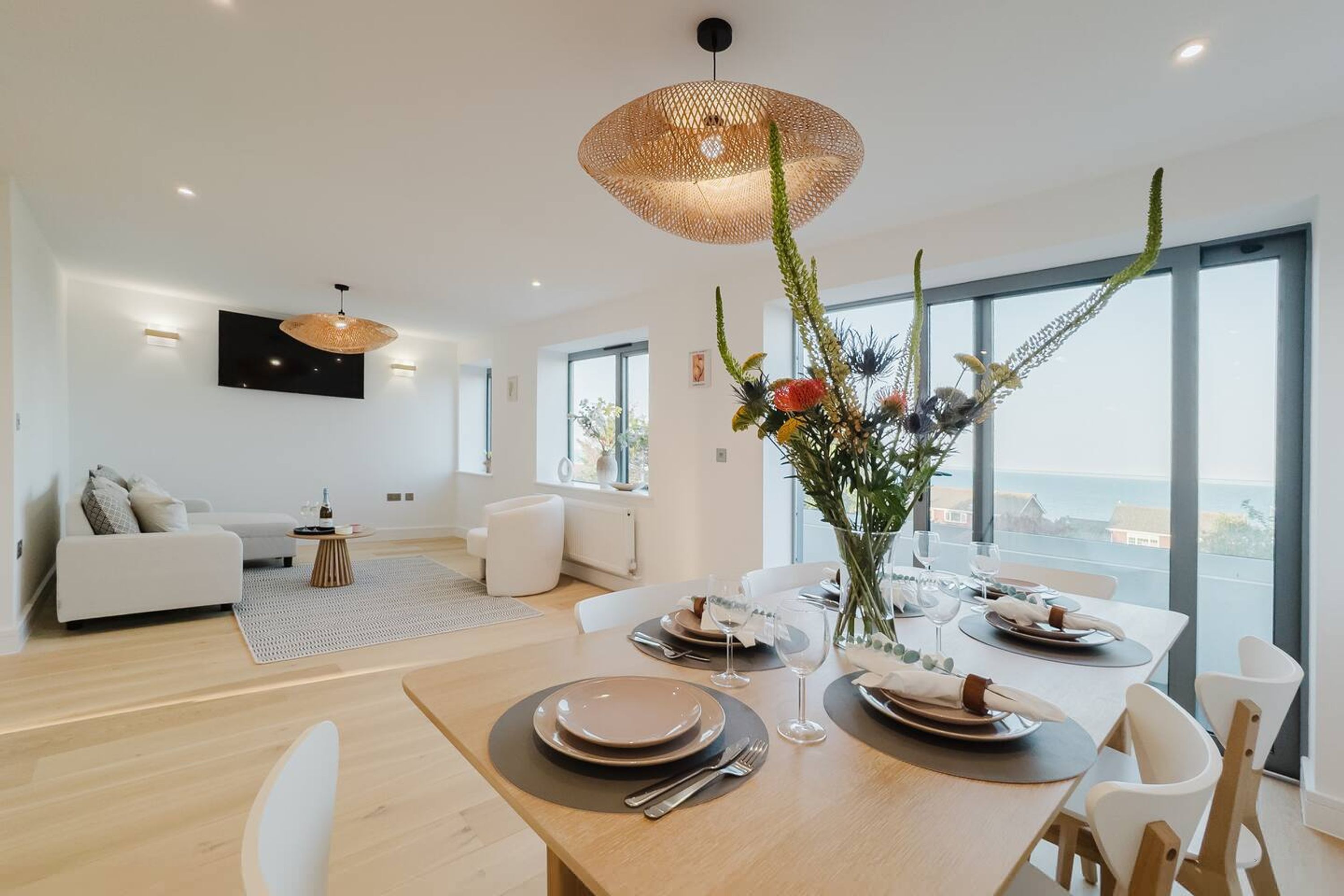 A modern dining area with a wooden table set for two, a large window with a view, and a living area with a sofa and a flat-screen TV.