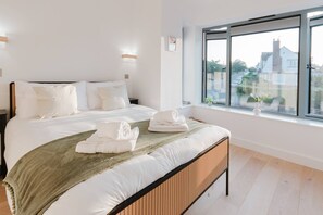 Ein ordentlich bezogenes Bett mit weißen Leinen und Handtüchern, ein Holz-Kopfteil und ein großes Fenster mit Blick auf Häuser.