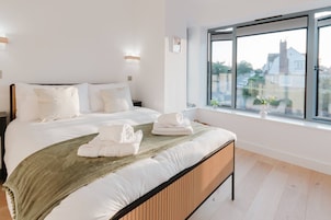 Cama bem arrumada com roupa de cama e toalhas brancas, cabeceira de madeira e uma janela grande com vista para casas.