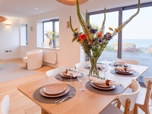 Un coin repas avec une table dressée pour quatre personnes, un vase de fleurs et une vue sur la mer par de grandes fenêtres.