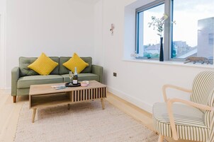 Un salón con un sofá verde, dos cojines amarillos, una mesa de centro de madera, un sillón a rayas y una ventana con vistas a la ciudad.