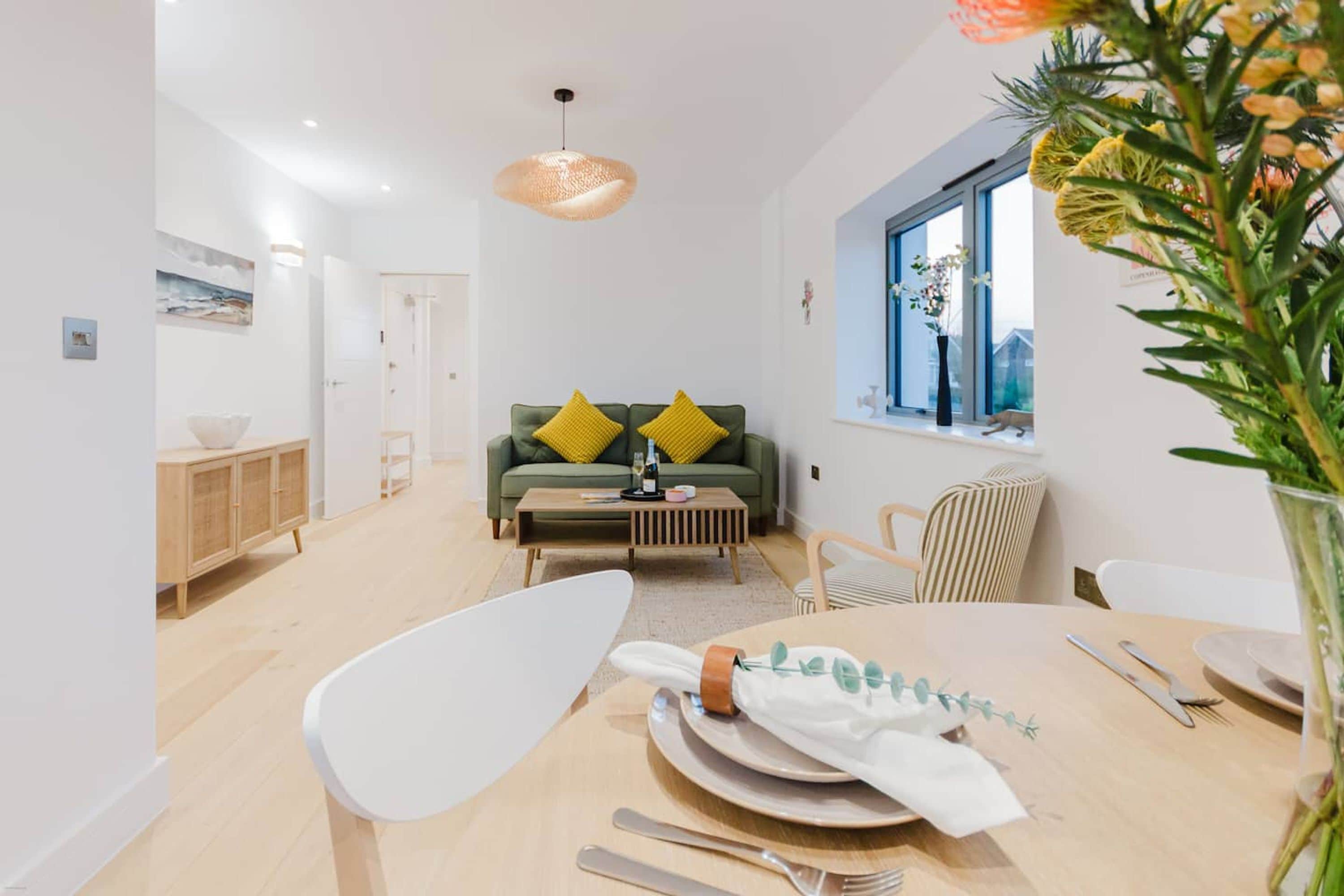 Ein moderner Essbereich mit einem Holztisch, gedeckt für eine Mahlzeit, einem grünen Sofa mit gelben Kissen und einem Fenster mit Blick auf Grünflächen.