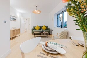 Un comedor moderno con una mesa de madera lista para una comida, un sofá verde con cojines amarillos, y una ventana con vistas a la vegetación.