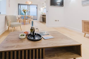 Uma sala de estar moderna com uma mesa de centro de madeira, uma garrafa de vinho e copos. Há uma poltrona listrada e uma mesa de jantar com cadeiras ao fundo.