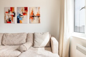 A living room with a sofa, a window with curtains, and three abstract paintings on the wall. - Bright Stepney Green City Stay (London)