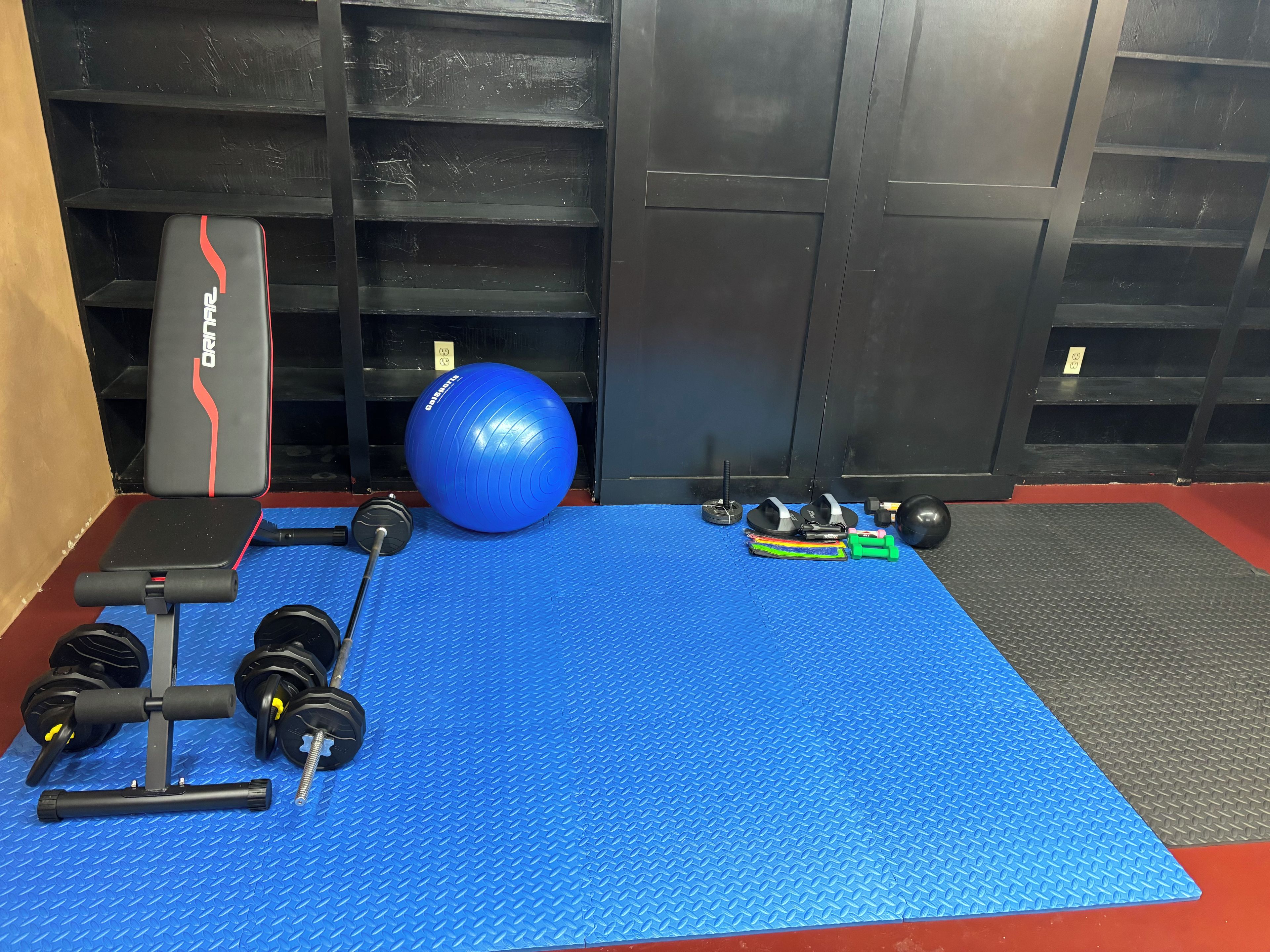 Une salle de sport équipée d’un tapis bleu, d’un banc de musculation, d’haltères, d’un ballon lesté et d’un support pour les exercices de squat.