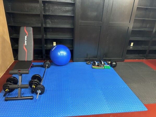 Un gimnasio con colchoneta azul, banco de pesas, mancuernas, pelota de medicina y una estructura para sentadillas.