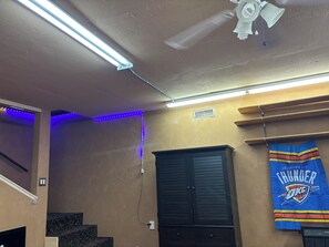 A room with a ceiling fan, fluorescent lighting, a blue LED strip, a black cabinet, and a wall-mounted Oklahoma City Thunder jersey.