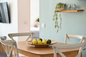 A dining area with a wooden table and chairs, a bowl of fruit, a hanging plant, and a shelf with decorative birds. - Bright Stay • Private Skyline Balcony by SeaNRent (Tel-Aviv)
