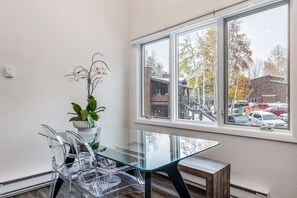 Una habitació amb taula de vidre, cadires transparents, una planta en test i una finestra amb vista a cotxes aparcats i edificis.