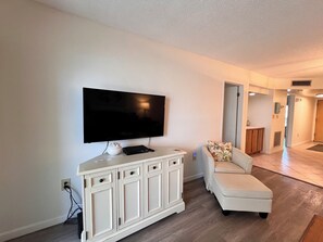 Sala de estar com televisão de tela plana na parede, um armário branco com gavetas, um sofá branco com almofada floral e piso de madeira.