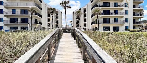 Une promenade en bois mène à un immeuble d’appartements moderne doté de nombreux balcons, avec un ciel dégagé en arrière-plan.