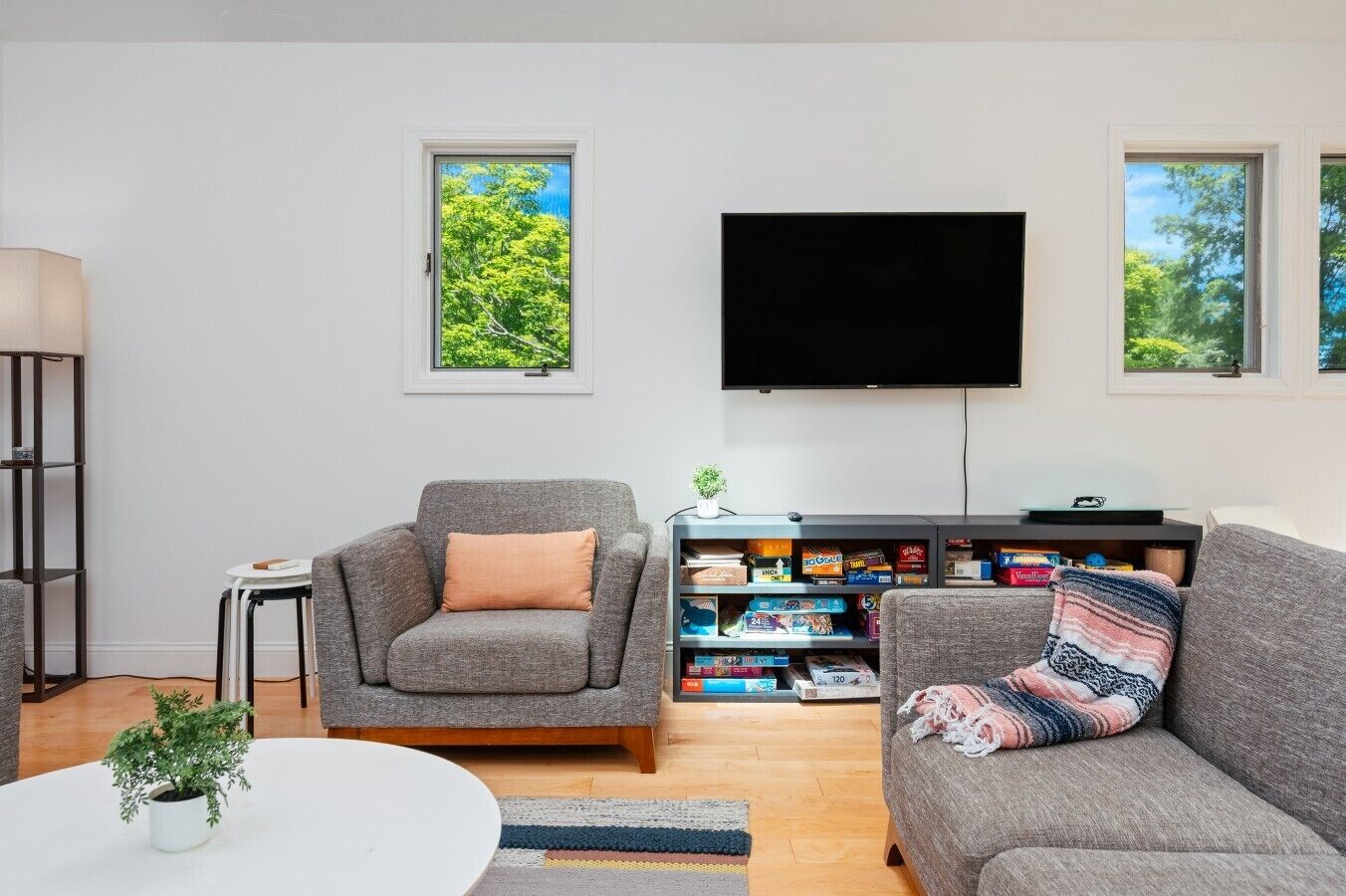 A living room with a grey sofa, a grey armchair, a flat-screen TV, a bookshelf with board games, and a window with a view of trees.