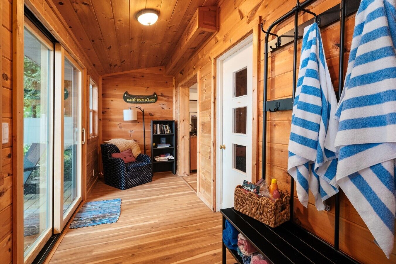 L’intérieur d’un chalet en bois, avec un canapé, une bibliothèque, une pancarte sur laquelle on peut lire « CAMP RUN-A-HURK », et des serviettes suspendues à un porte-serviettes.