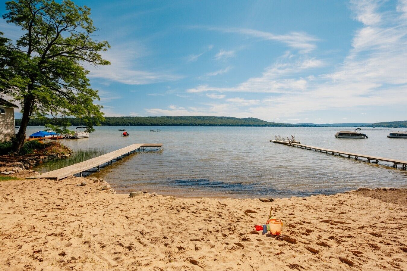 A sandy beach with a wooden dock extending into calm water, surrounded by trees and boats.