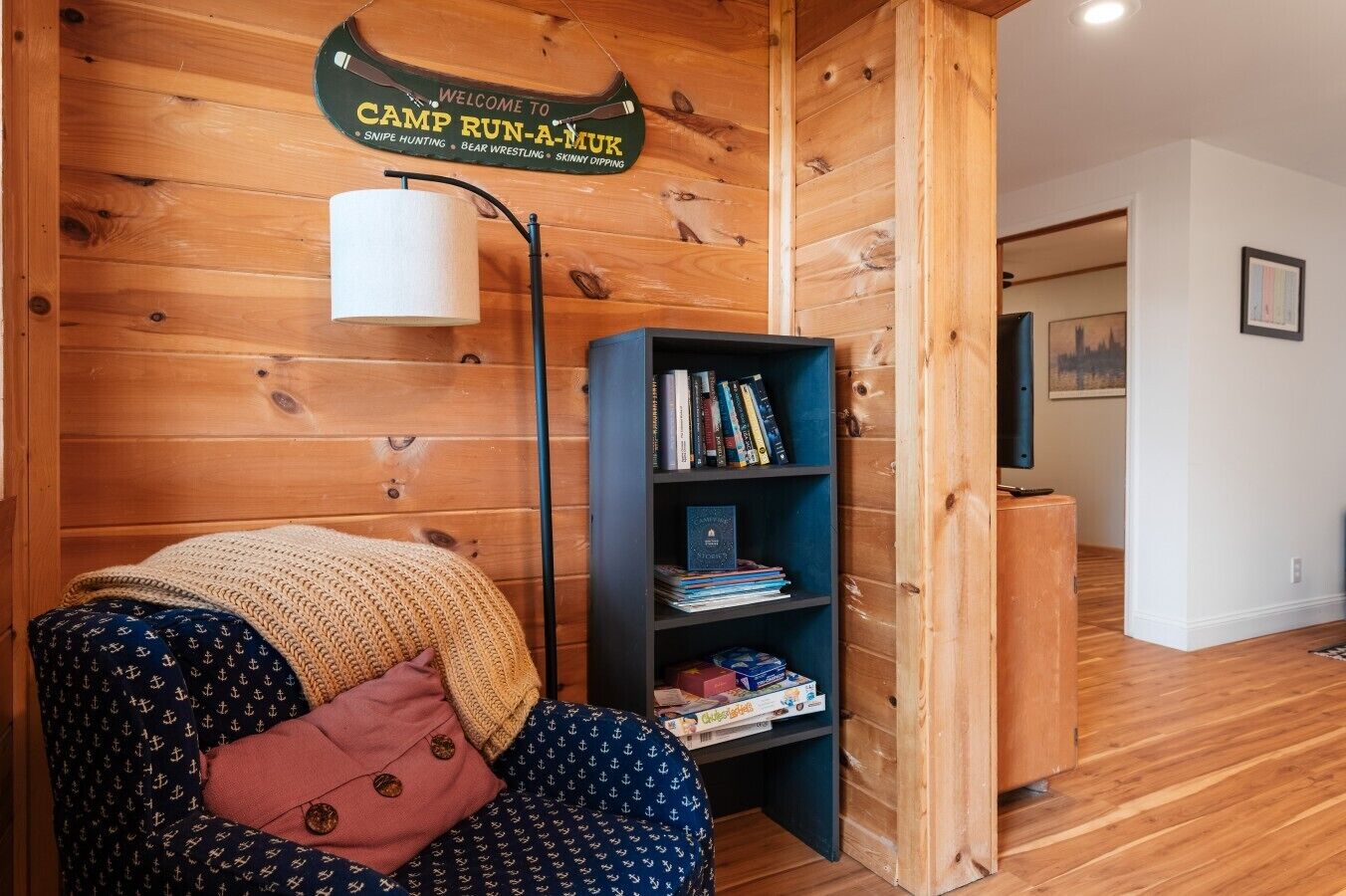 A cozy room with a wooden wall, a bookshelf, a chair with a blanket, and a lamp.