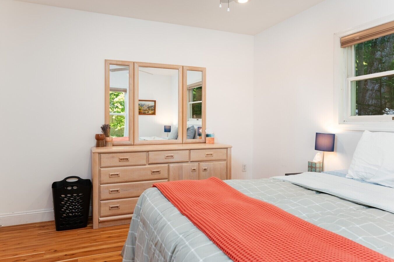 A bedroom with a bed, a dresser with a mirror, a window, and a nightstand with a lamp.