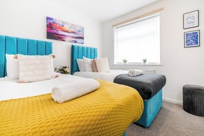 Ein Hotelzimmer mit einem großen Bett, einem kleineren Bett und einem Schlafsofa. Es gibt ein Fenster mit Jalousien, ein Bild an der Wand und eine kleine Pflanze auf der Fensterbank.